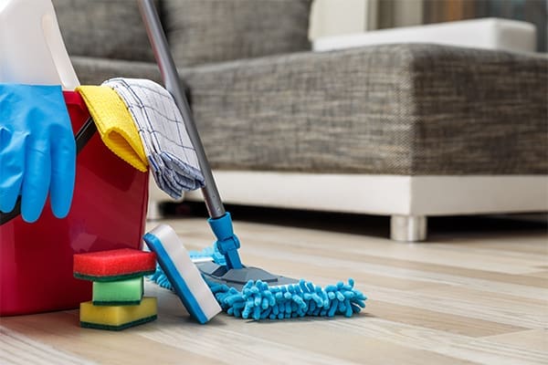 A mop stands beside a cleaning bucket, sponges, rubber gloves and cleaning cloths. A brown and white couch is in the background.