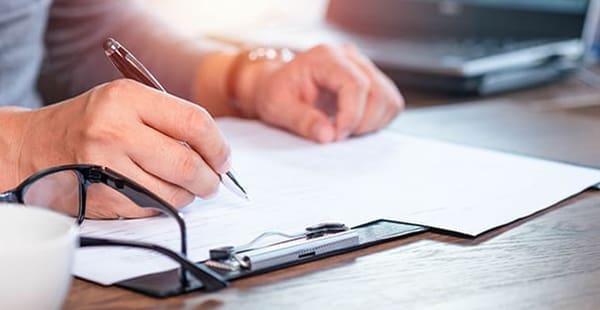 A photo of a hand holding a pen on a sheet of paper attached to a clipboard. The person is writing questions to ask their mesothelioma doctor.