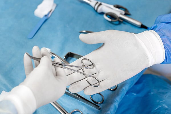 Healthcare providers in sterile garb and gloves exchange a surgical instrument in the operating room. This exchange is emblematic of the potential rise in surgery-based mesothelioma treatments among Medicare patients.