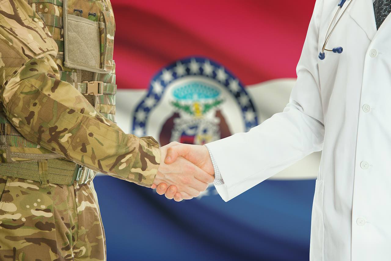 Soldier in uniform shaking hands with doctor with Missouri state flag in background