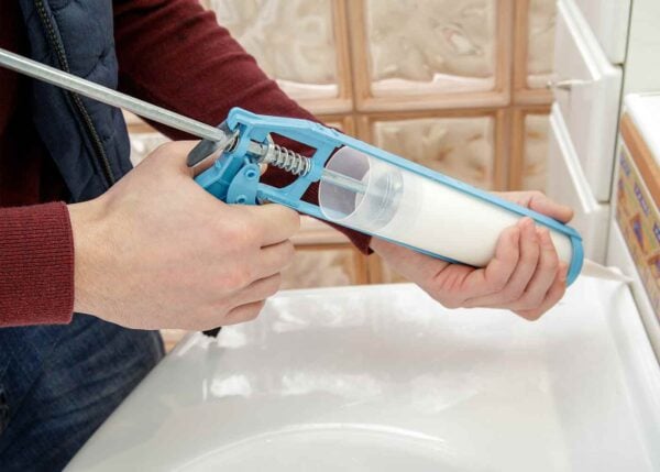 Image shows a person holding a contractor-style sealant gun in both hands, using the gun to apply sealant around the edge of a white porcelain vanity.