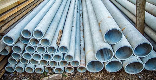 Cement asbestos pipes stacked up in a lumberyard. The pipes are a light blue-gray color with rust and other visible signs of deterioration