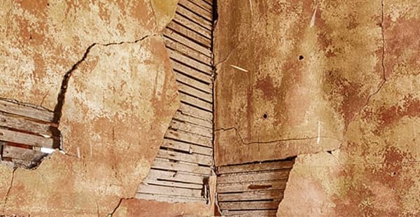 Photograph showing what asbestos plaster walls look like. It shows the junction between two walls composed of lath and plaster. Plaster has fallen off of some areas of the wall, exposing the lath layer underneath.