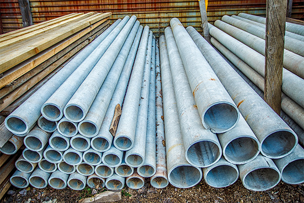 Cement asbestos pipes stacked up in a lumberyard. The pipes are a light blue-gray color with rust and other visible signs of deterioration