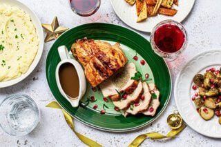 A holiday meal featuring sliced turkey on a green plate with gravy in a white gravy boat. A bowl of mashed potatoes, a plate of roast vegetables, a glass of water and glasses of red wine are also seen. Gold ribbons and stars decorate the place setting.