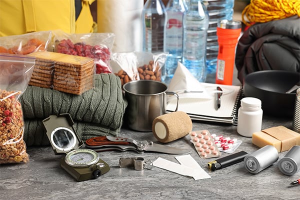 Image depicts the scattered components of a disaster supply kit, including a compass, medications, mylar blankets, shelf-stable foods and water.