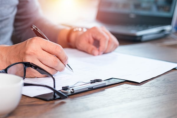 A photo of a hand holding a pen on a sheet of paper attached to a clipboard. The person is writing questions to ask their mesothelioma doctor.