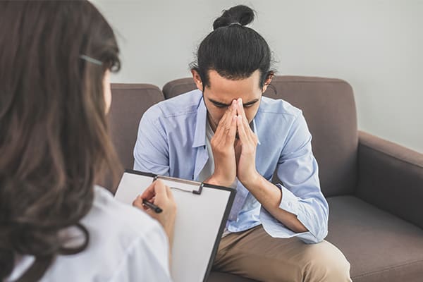 A young man sits with his head in hands in stress, while a doctor takes notes on a clipboard.