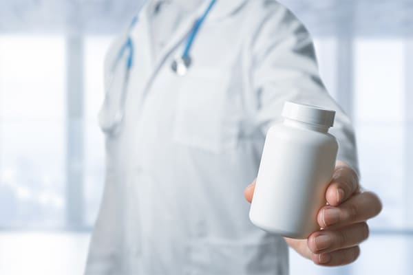 A doctor wearing a white coat with a blue stethoscope draped around the collar holds out a bottle of orphan drug pills.