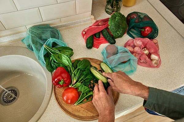 A pair of hands slicing a cucumber next to a sink. Other fresh produce is around the hands, including red peppers, cucumbers, tomatoes, mushrooms, basil and broccoli.