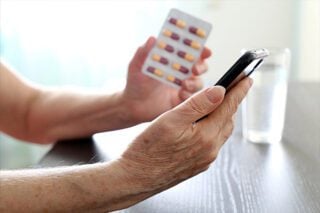 A picture of a mesothelioma patient's hand holding a smartphone to use an app to track his medications he holds in his other hand.
