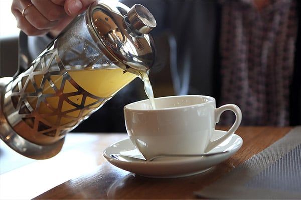 A photo showing yellowish-colored tea being poured from a glass and metal teapot into a white teacup.