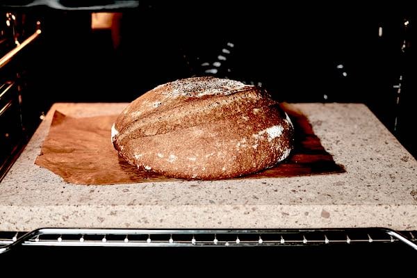 A loaf of home-baked bread sits atop parchment paper on a regular baking stone, all on the middle rack of a consumer-grade oven. This setup demonstrates an expert-recommended breadmaking method using a regular baking stone, not anything made of asbestos.